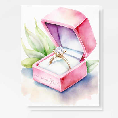 A beautiful watercolor engagement card featuring a gold diamond ring inside a pink velvet box with green botanical accents. The words "She Said Yes!" are delicately written on the box.