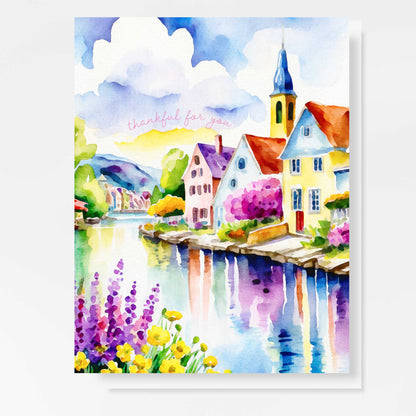 Watercolor greeting card featuring a charming riverside village with colorful houses, a church steeple, blooming flowers, and a reflective river. The words "thankful for you" are written in soft script. A2 size (5.5" x 4.25") with a white envelope.