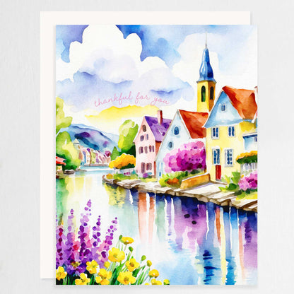 Watercolor greeting card featuring a charming riverside village with colorful houses, a church steeple, blooming flowers, and a reflective river. The words "thankful for you" are written in soft script. A2 size (5.5" x 4.25") with a white envelope.