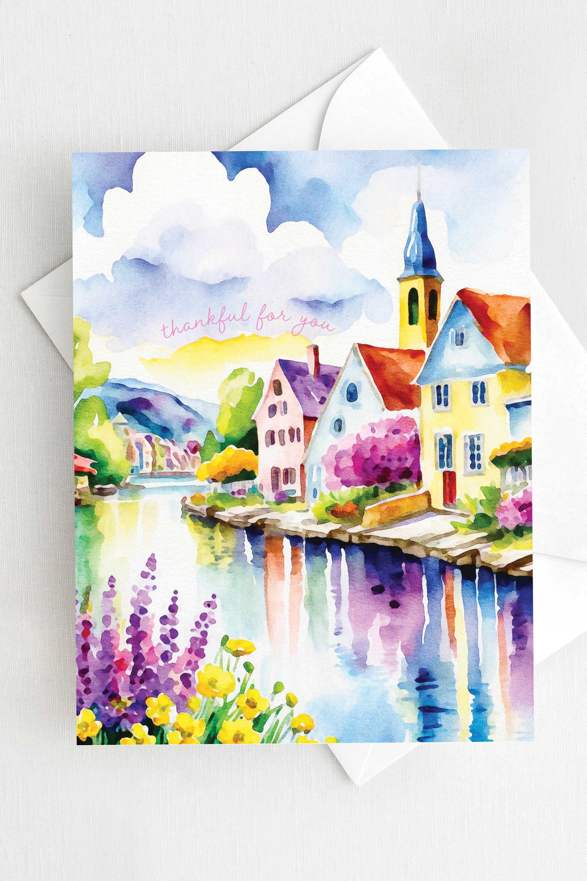 Watercolor greeting card featuring a charming riverside village with colorful houses, a church steeple, blooming flowers, and a reflective river. The words "thankful for you" are written in soft script. A2 size (5.5" x 4.25") with a white envelope.