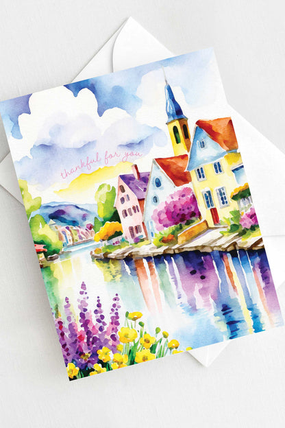 Watercolor greeting card featuring a charming riverside village with colorful houses, a church steeple, blooming flowers, and a reflective river. The words "thankful for you" are written in soft script. A2 size (5.5" x 4.25") with a white envelope.