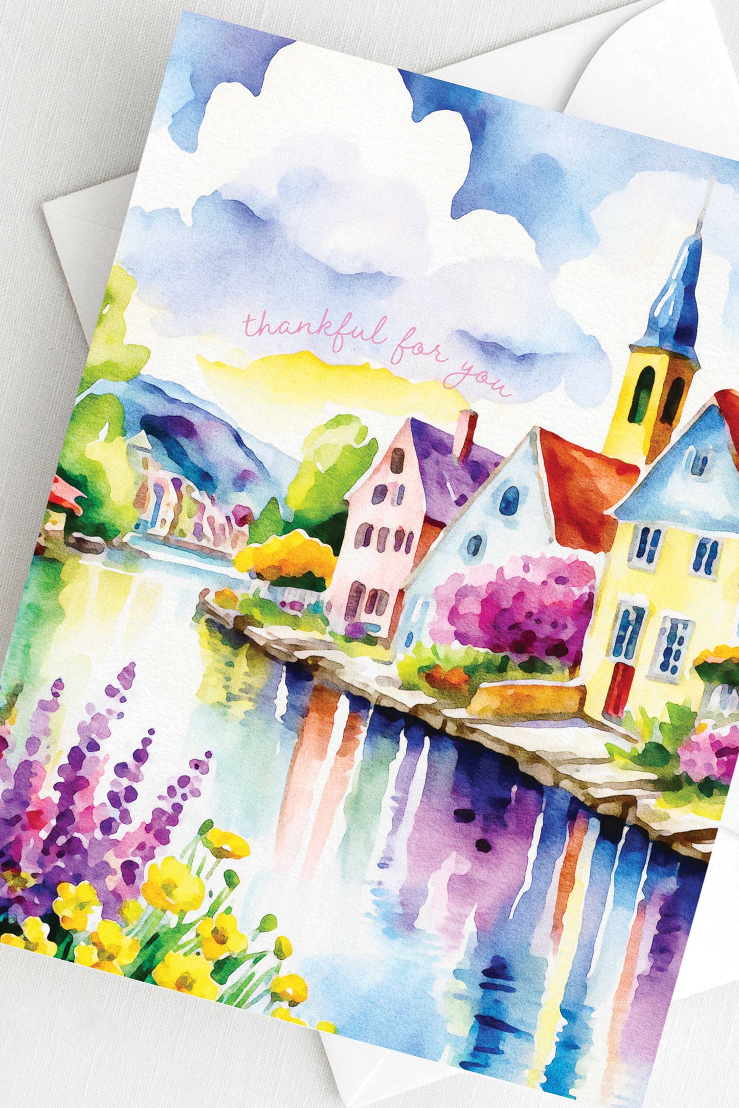 Watercolor greeting card featuring a charming riverside village with colorful houses, a church steeple, blooming flowers, and a reflective river. The words "thankful for you" are written in soft script. A2 size (5.5" x 4.25") with a white envelope.