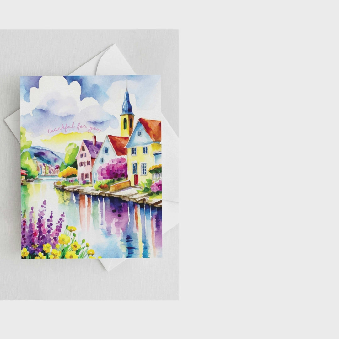 Watercolor greeting card featuring a charming riverside village with colorful houses, a church steeple, blooming flowers, and a reflective river. The words "thankful for you" are written in soft script. A2 size (5.5" x 4.25") with a white envelope.